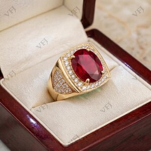 May include: A gold ring featuring a large, oval-cut deep red gemstone at the center, surrounded by a halo of small, clear, round-cut stones. The band is wide and also encrusted with multiple small, clear, round-cut stones along the sides.