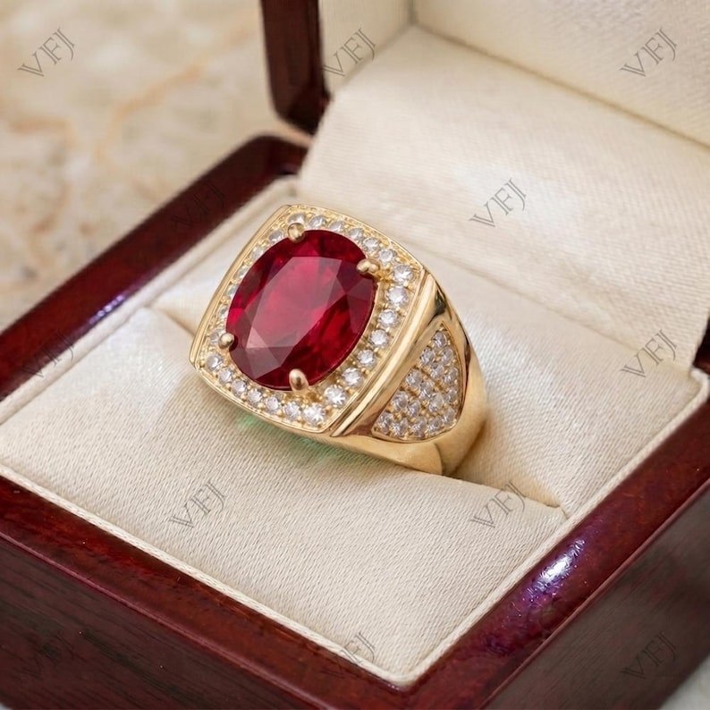 May include: A gold ring featuring a large, oval-cut, deep red gemstone at the center, surrounded by a halo of small, clear, round-cut stones. The band is also adorned with rows of small, clear, round-cut stones along the sides of the setting.