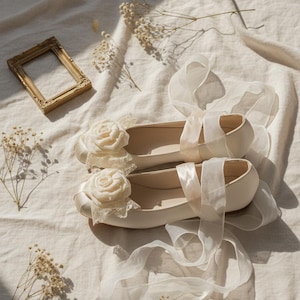 Puede incluir: Un par de bailarinas color marfil con adornos de rosas y lazos de organza. Sobre tela crema, con un pequeño marco dorado y flores secas. Zapatos de estilo delicado y femenino, ideales para ocasiones especiales. Miden unos 24 cm.