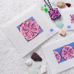 May include: Three needlepoint canvases and a small stitched ornament featuring pink and purple sand dollar designs on blue backgrounds. The canvases include small purple labels with the text Let's Stitch Studio, accompanied by various pieces of sea glass.