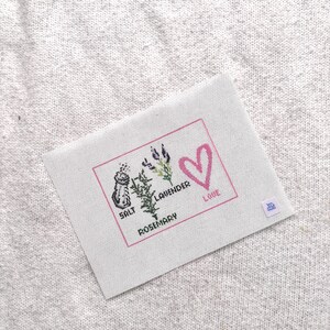 May include: A rectangular white fabric piece featuring a cross-stitch design enclosed in a pink border. The design includes a salt shaker, sprigs of lavender and rosemary, and a pink heart with the text SALT, LAVENDER, ROSEMARY, and LOVE.