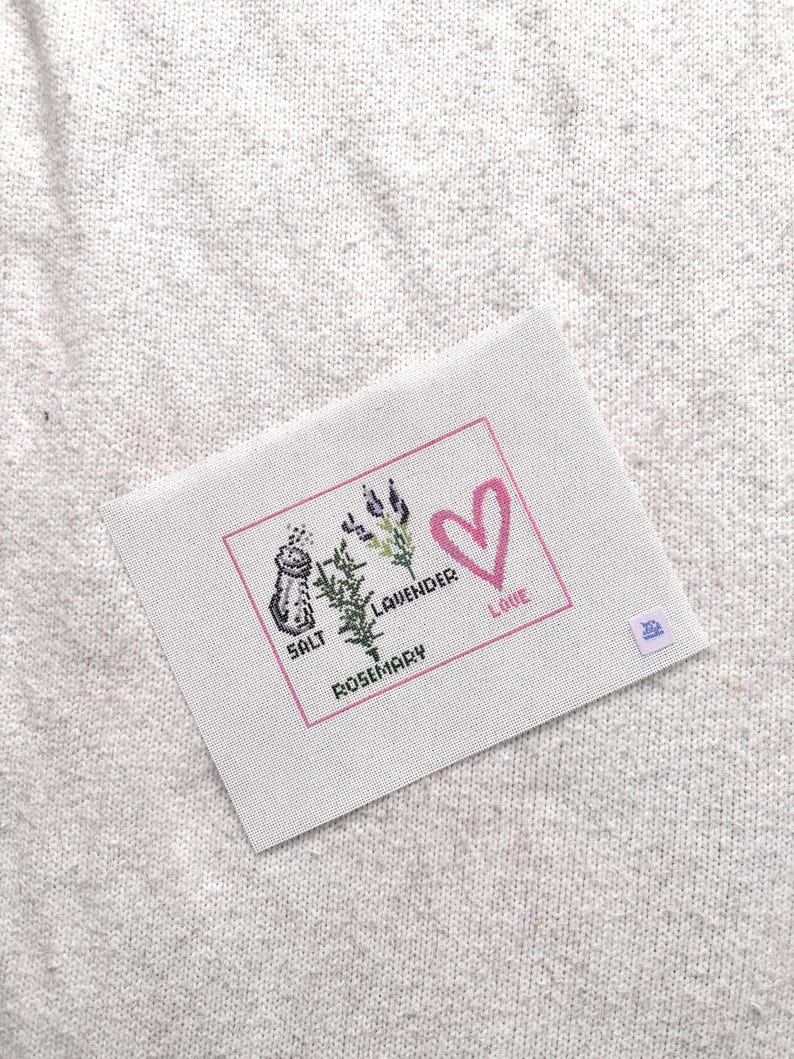 May include: A rectangular white fabric piece featuring a cross-stitch design enclosed in a pink border. The design includes a salt shaker, sprigs of lavender and rosemary, and a pink heart with the text SALT, LAVENDER, ROSEMARY, and LOVE.
