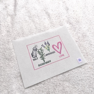 May include: A rectangular white needlepoint canvas featuring a pink border and cross-stitched designs of a salt shaker, a sprig of rosemary, and lavender. The text reads SALT, ROSEMARY, LAVENDER, and LOVE, accompanied by a pink heart outline.