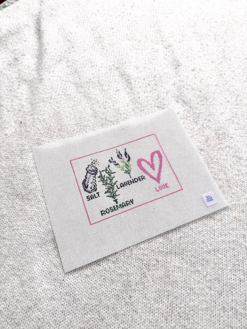 May include: A rectangular white needlepoint canvas featuring a pink border and cross-stitched designs of a salt shaker, a sprig of rosemary, and lavender. The text reads SALT, ROSEMARY, LAVENDER, and LOVE, accompanied by a pink heart outline.