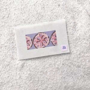 Könnte beinhalten: Eine rechteckige Stickleinwand mit einem sich wiederholenden Muster aus drei rosa und violetten Sanddollars auf hellblauem Grund. Ein kleines quadratisches Etikett in der unteren rechten Ecke zeigt den Text Let's Stitch Studio in Violett.