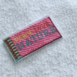 May include: A rectangular, pink needlepoint patch with the words "PEACE LOVE NATURE" in yellow and teal. The patch has a light blue border and a green, red, and brown striped edge. The patch is set against a white, textured background.