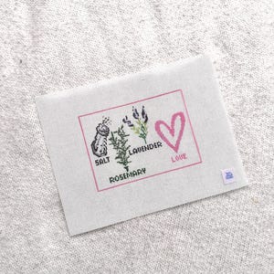 May include: A rectangular white fabric piece featuring a cross-stitch design enclosed in a pink border. The design includes a salt shaker, a rosemary sprig, lavender stalks, and a pink heart, with the text SALT, ROSEMARY, LAVENDER, and LOVE.