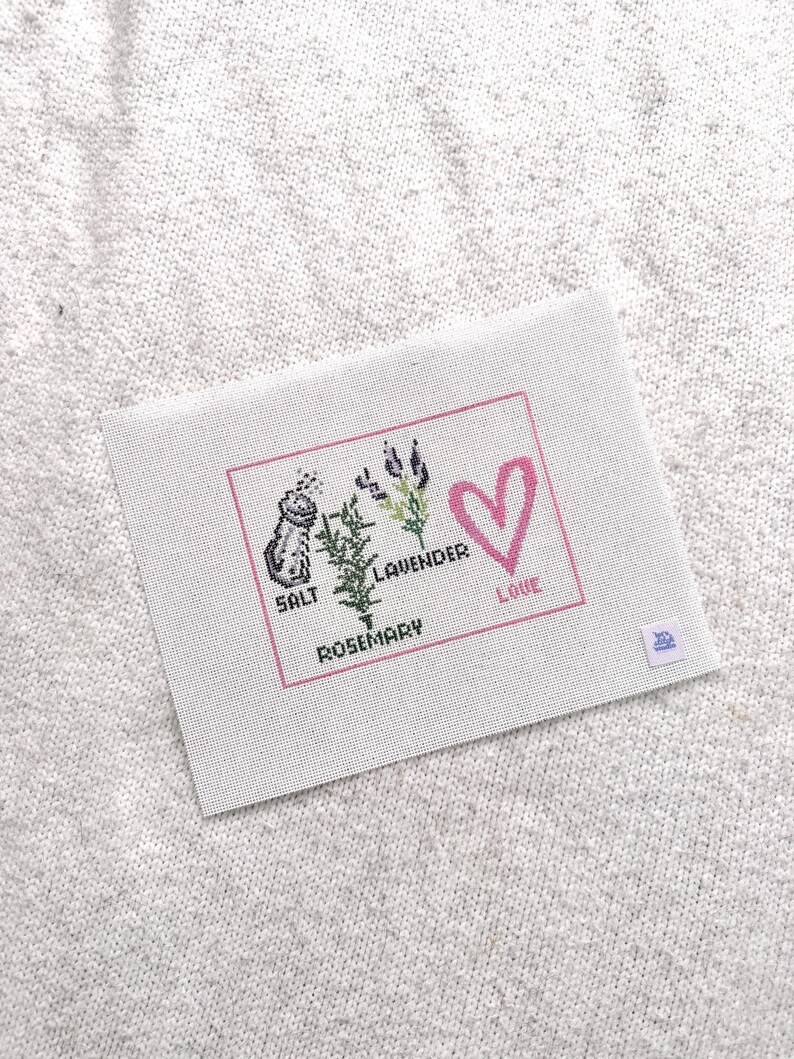 May include: A rectangular white fabric piece featuring a cross-stitch design enclosed in a pink border. The design includes a salt shaker, a rosemary sprig, lavender stalks, and a pink heart, with the text SALT, ROSEMARY, LAVENDER, and LOVE.