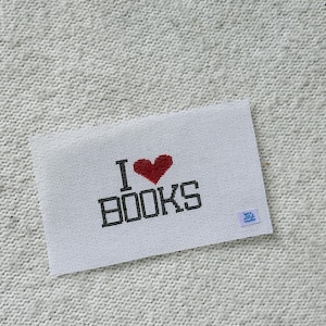 May include: White needlepoint canvas with the text "I ❤️ BOOKS" in black and red. The heart is red, and the word BOOKS is in a bold, blocky font. A small blue tag is in the bottom right corner.