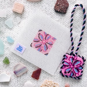 Puede incluir: Un lienzo cuadrado de punto de aguja blanco con un patrón floral geométrico rosa y azul y una pequeña etiqueta azul con la inscripción Let's Stitch Studio, junto a un adorno cuadrado terminado con un patrón a juego y un cordón trenzado negro, rosa y azul.