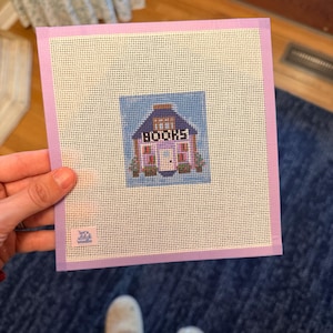 May include: A square needlepoint canvas featuring a central design of a small building with a purple roof and walls, with the word BOOKS printed in black letters above the door. The canvas has a light purple border and a small logo in the corner.