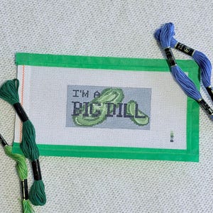 May include: A needlepoint canvas with the text "I'M A PIC DILL" stitched in green and black. The canvas is bordered in green and surrounded by skeins of blue and green embroidery floss.
