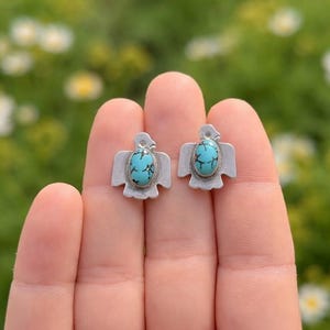 May include: A pair of silver-toned stud earrings, each featuring a turquoise stone set within a stylized bird design. The earrings are displayed on a hand against a blurred green and white floral background.