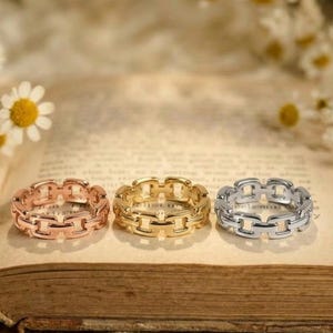 May include: Three chain-link style rings in rose gold, gold, and silver, displayed on an open book. The rings are the focal point, with daisies and other flowers in the background. The rings are a jewellery item.