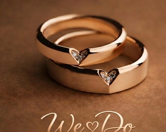 May include: Two rose gold wedding rings with a heart-shaped design, each embedded with small diamonds. The rings are stacked on top of each other, with the words "We Do" in a cursive font at the bottom.