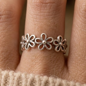Puede incluir: Un anillo de plata con un diseño floral repetido de flores tipo margarita. El anillo se lleva en un dedo, mostrando los delicados detalles de los pétalos y la artesanía general. El anillo es una pieza de joyería.