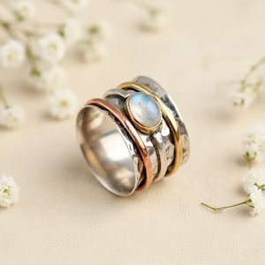 Puede incluir: Un anillo giratorio de plata con una piedra lunar ovalada en el centro. El anillo presenta bandas de plata, cobre y oro. La piedra lunar está engastada en un bisel dorado. El anillo se muestra sobre un fondo neutro con flores blancas.