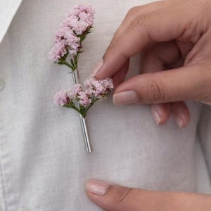 Op de afbeelding: Een zilveren reversspeld met een takje kleine, lichtroze bloemen. De speld wordt op een wit overhemd geplaatst. De bloemen hebben groene stelen en zijn gegroepeerd. Een stijlvol accessoire.