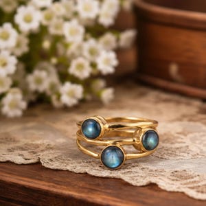May include: A stackable gold ring set with three rings, each featuring a round, blue gemstone. The rings are displayed on a lace doily and a wooden surface, with white flowers in the background.