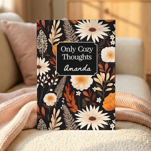 May include: A rectangular notebook with a black cover featuring a floral pattern of white, orange, and brown flowers and leaves. A central black rectangular label displays the white text "Only Cozy Thoughts" above the name "Amanda" in white script.