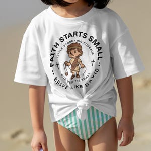 May include: White t-shirt with a circular graphic featuring a cartoon illustration of a boy holding a sling. The graphic includes the text "Faith Starts Small... Brave Like David." The shirt is knotted at the waist and worn with striped swimwear.