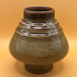 May include: A ceramic vase with a bulbous body and a flared rim. The vase is primarily olive green with bands of brown and reddish-brown around the neck. The base is a darker brown. A decorative piece for the home.