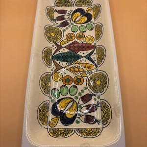 Może przedstawiać: Prostokątna ceramiczna taca do serwowania w kolorze kremowym z białym obramowaniem. Taca posiada kolorowy wzór ryb, cytryn, oliwek i innych elementów dekoracyjnych w kolorze żółtym, pomarańczowym, zielonym i niebieskim.