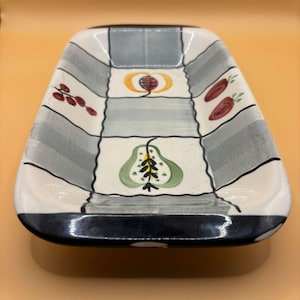 Może przedstawiać: Dekoracyjny ceramiczny talerz do serwowania o prostokątnym kształcie i zaokrąglonych rogach. Talerz posiada wzór w szachownicę w kolorze szarym i białym, z namalowanymi wzorami owoców i warzyw w kolorze czerwonym, żółtym i zielonym. Obręcz jest granatowa.