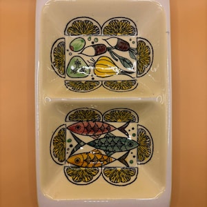 Może przedstawiać: Prostokątny, dzielony ceramiczny talerz do serwowania z białą obwódką i jasnożółtym wnętrzem. Talerz jest ozdobiony ręcznie malowanymi obrazami ryb i warzyw, otoczonych plasterkami cytryny.