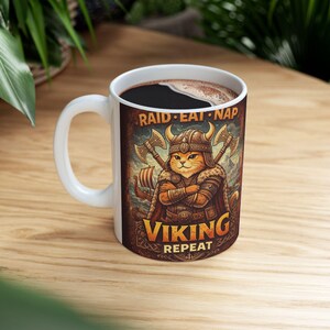 Puede incluir: Taza de cerámica blanca con café negro. La taza presenta una ilustración de dibujos animados de un gato vikingo con hachas cruzadas y el texto "RAID EAT NAP VIKING REPEAT".
