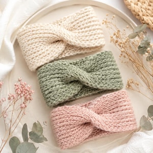 Könnte beinhalten: Drei gestrickte Stirnbänder mit einem gedrehten Design auf der Vorderseite. Die Stirnbänder sind in den Farben Creme, Salbeigrün und zartem Rosa gehalten. Jedes Stück weist eine gleichmäßige Rippstrickstruktur auf, die ein einheitliches Bild ergibt.