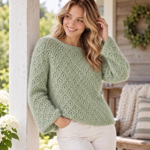 May include: A sage green long-sleeved sweater featuring a textured diamond-shaped openwork knit pattern, a round neckline, and ribbed cuffs. The garment is paired with white trousers, creating a neutral and soft aesthetic for the ensemble.