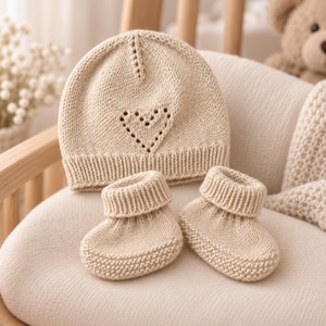 Pode incluir: Um gorro de malha bege para bebé e um par de botinhas a combinar. O gorro apresenta uma borda canelada e um padrão de orifícios em forma de coração na frente. As botinhas têm um punho canelado e um design de malha texturizada no pé.