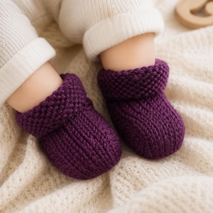 May include: A pair of deep purple knitted baby booties featuring a textured garter stitch cuff and a ribbed knit foot section. The booties are worn over white ribbed leggings, showcasing a thick, soft yarn construction with a folded top edge.