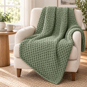 Puede incluir: Una manta de color verde salvia con una textura tejida gruesa tipo cesta, colocada sobre un sillón tapizado de color crema. La manta presenta bucles de hilo gruesos e interconectados que crean un patrón uniforme de rejilla en la tela.