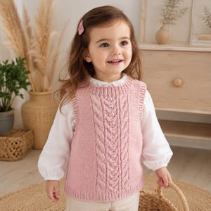 May include: A light pink sleeveless knitted sweater vest featuring a vertical cable knit pattern on the front, a ribbed crew neckline, and ribbed armholes. The garment is worn over a white long-sleeved shirt with a high collar and light-coloured trousers.
