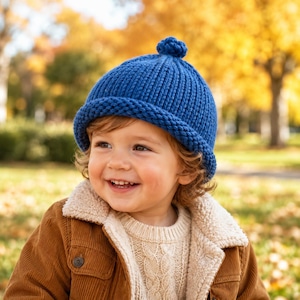 Puede incluir: Un gorro de punto azul vibrante con una textura acanalada gruesa, un borde ancho doblado y un pequeño pompón en la corona. El gorro está confeccionado con hilo grueso y ofrece un ajuste ceñido, mostrando un diseño clásico y tejido.