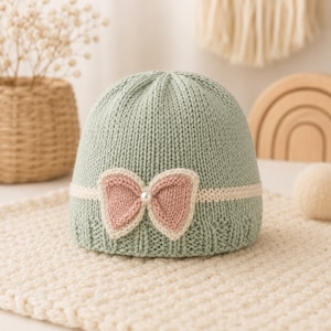 Pode incluir: Um gorro de malha verde-sálvia com uma faixa horizontal de cor creme ao centro. Um laço de malha rosa suave está preso na parte frontal da faixa, acentuado com uma pequena pérola branca no centro. O gorro apresenta uma textura canelada.