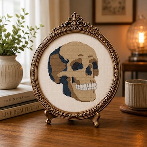 May include: A circular cross-stitch embroidery featuring a tan and dark blue skull design on a cream-colored fabric background, encased in an ornate, antique-style gold-toned metal frame with a decorative top crest and a small stand.