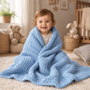 Può includere: Una coperta in maglia grossa color azzurro avvolta attorno a un bambino sorridente. La coperta presenta una struttura in filato spesso e testurizzato con un motivo a punti coerente, offrendo un aspetto morbido e accogliente sulle spalle.