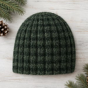 May include: A dark forest green beanie hat featuring a vertical ribbed knit pattern. The hat is constructed from a variegated yarn that blends deep green tones with subtle lighter green highlights throughout the textured fabric surface.