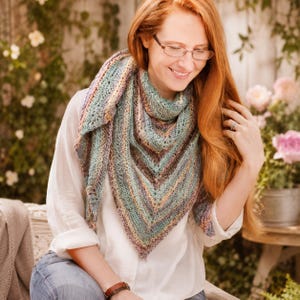 May include: A triangular knit shawl featuring a variegated color palette of teal, soft green, and muted orange tones. The accessory displays a textured stitch pattern with a pointed bottom edge, draped over a white long-sleeve shirt and blue jeans.