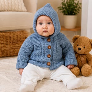 May include: A baby wearing a dusty blue knitted hooded cardigan with a textured cable knit pattern and four round wooden buttons down the front. The cardigan features long sleeves with ribbed cuffs and a pointed hood, paired with plain white pants.