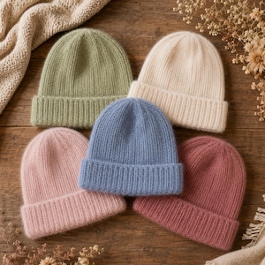 Può includere: Una collezione di cinque berretti in maglia in vari colori solidi, tra cui verde salvia, crema, blu polvere, rosa chiaro e malva. Ogni berretto presenta una consistenza a coste, un risvolto piegato alla base e un aspetto morbido e soffice.