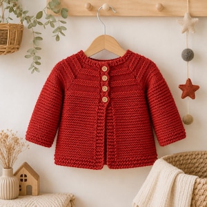 May include: A long-sleeved, deep red knitted cardigan featuring a round neckline and a vertical row of four light-colored wooden buttons down the center front. The garment is crafted with a textured garter stitch pattern throughout the body and sleeves.