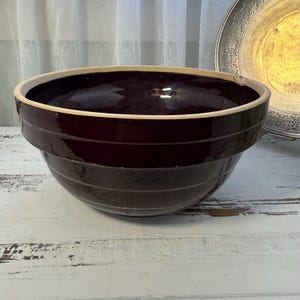 Puede incluir: Un gran cuenco de cerámica marrón oscuro con un borde de color crema. El cuenco tiene estrías horizontales alrededor del exterior y se apoya sobre una superficie de madera blanca. En el fondo se ve un plato decorativo de plata.