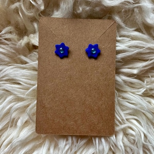 Könnte beinhalten: Ein Paar blaue, blütenförmige Ohrstecker mit einem kleinen, durchsichtigen Edelstein in der Mitte. Die Ohrringe werden auf einer braunen Karte vor einem flauschigen, weißen Hintergrund präsentiert.