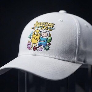 May include: A white baseball cap featuring a colorful graphic print of the Adventure Time logo with characters Jake the Dog and Finn the Human. The design includes various small animated figures surrounding the central characters on the front panel.