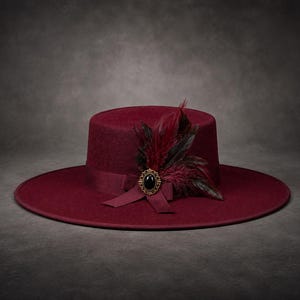 Pode incluir: Um chapéu de aba larga em bordô profundo com copa plana e fita a condizer. O chapéu é adornado com um grupo de penas vermelho-escuras e pretas e um broche oval dourado com uma pedra preta lisa no centro.