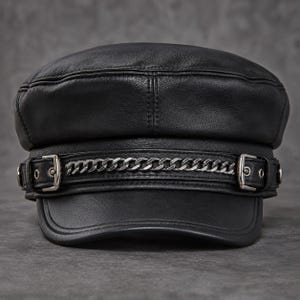 Puede incluir: Una gorra de estilo marinero en cuero negro con una corona estructurada y una visera corta y curva. Una tira de cuero horizontal adornada con una cadena de metal plateado y dos pequeñas hebillas plateadas se sitúa en la parte frontal.